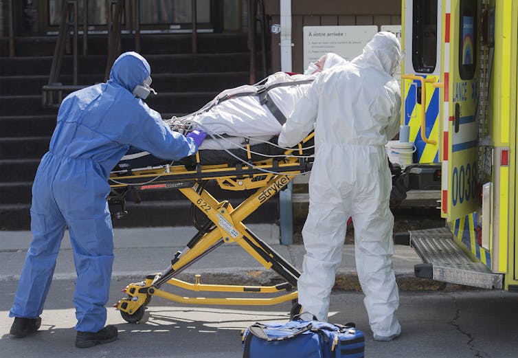 Paramedics in protective equipment wheel a patient wrapped in blankets and strapped onto a stretcher toward a waiting ambulance.