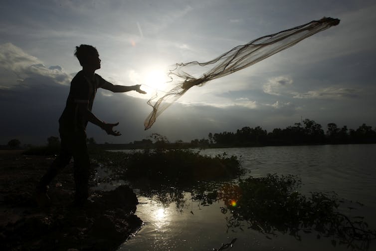 A man tosses a fishing net into a body of water with the sun rising in the background.