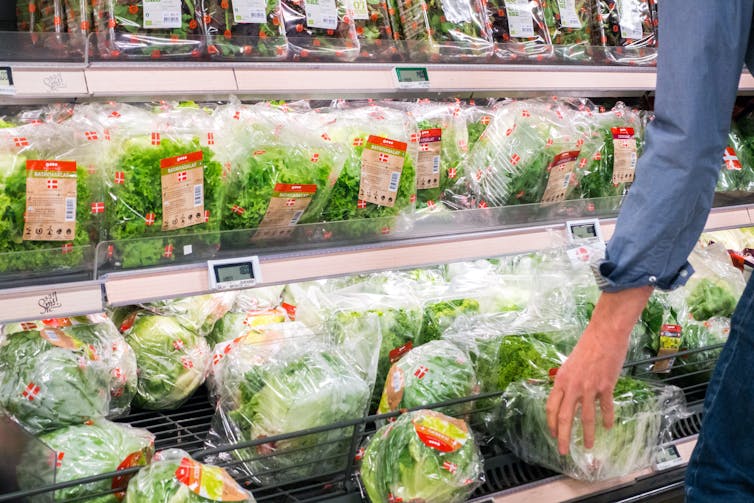 A person's arm reaches for a package of greens from a selection of greens and lettuces in a grocery store.