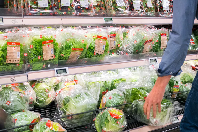 A person's arm reaches for a package of greens from a selction of greens and lettuces in a grocery story.