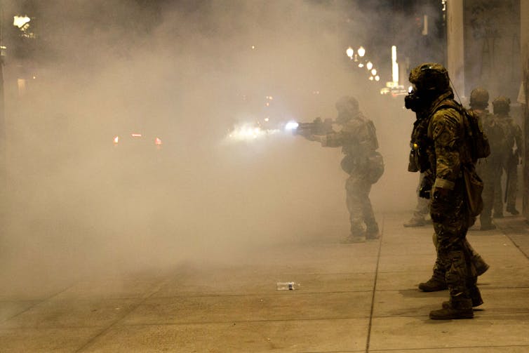 A federal officer in a heavy camouflage uniform fires into a cloud of smoke