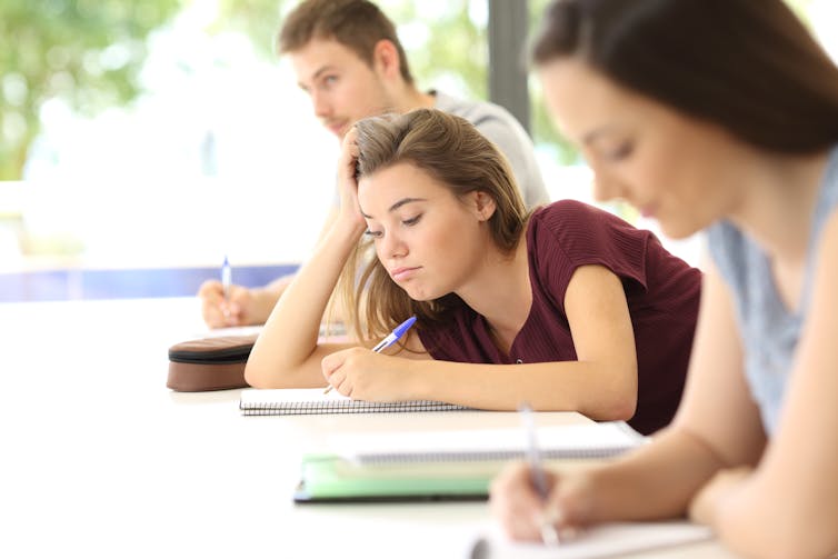 A female student taking notes at her desk but looking bored.