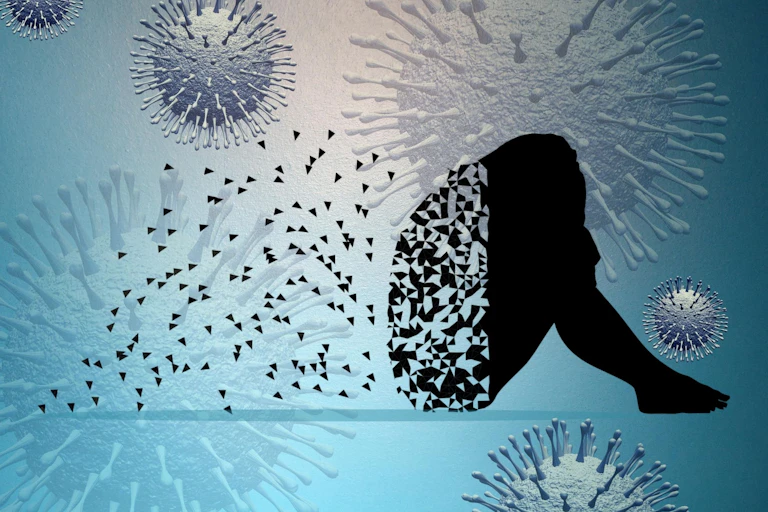 Silhouette of a person sitting with their head on their knees. The silhouette is starting to disintegrate and pieces of the silhouette float away in a blue background of coronaviruses.