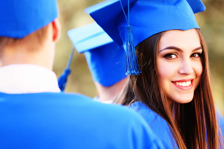 Female university graduate in blue hat and gown looking over her shoulder at camera.