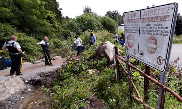 Police officers direct a group of migrant walking along a rocky path, with trees in the background and a warning sign to the right of the scene.