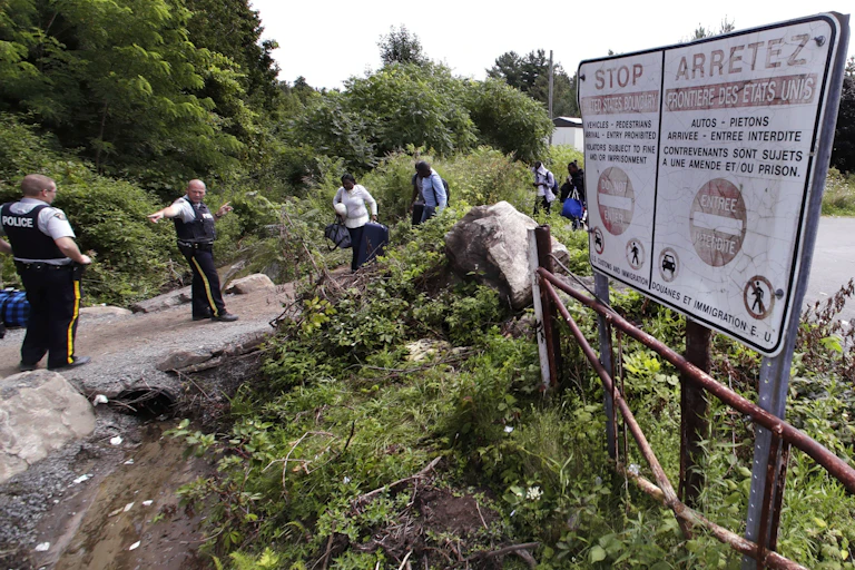 Police officers direct a group of migrant walking along a rocky path, with trees in the background and a warning sign to the right of the scene.