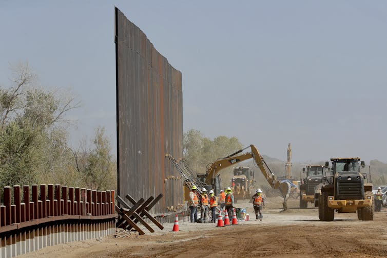A tall portion of metal wall stands as construction workers work at its base, with a bulldozer and tractors in the background.