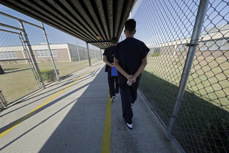 Men are seen walking in an outdoor, caged walkway with their arms behind their backs and dressed in correctional facility garb.