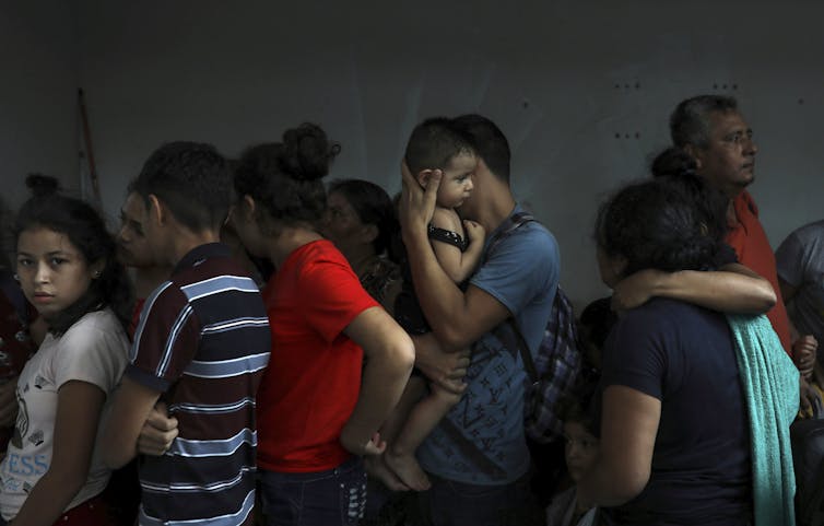 A group of migrants, including a man holding a small baby, are seen standing together in a darkened room. One teenaged girl looks directly at the camera.