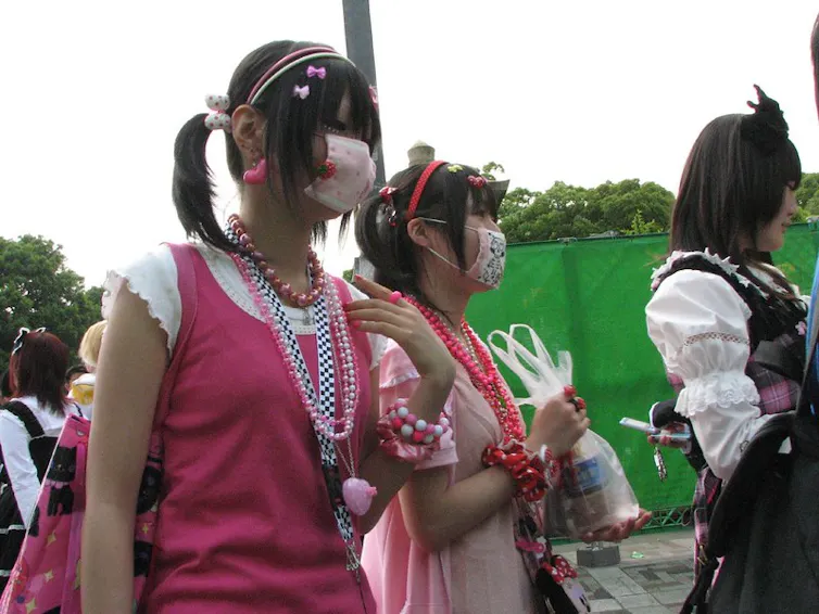 Two Japanese Harajuku girls in pink dresses wear face masks.