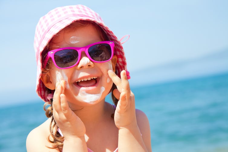 Young girl rubbing sunscreen on her face, wearing a bucket hat and sunglasses.