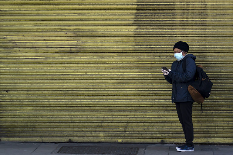 A woman in mask stands in front of a closed shop with its shutters down