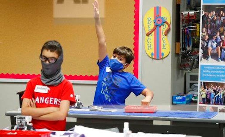Two boys in a classroom wearing masks, with one raising his hand.