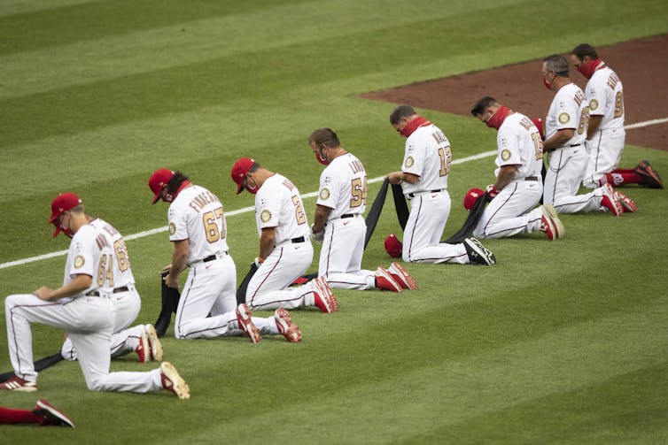 A row of baseball players, each holding the same long strip of black cloth, kneel.