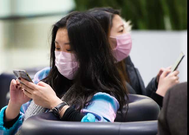 Two Asian women wearing face masks sit on couches while using their phones. 