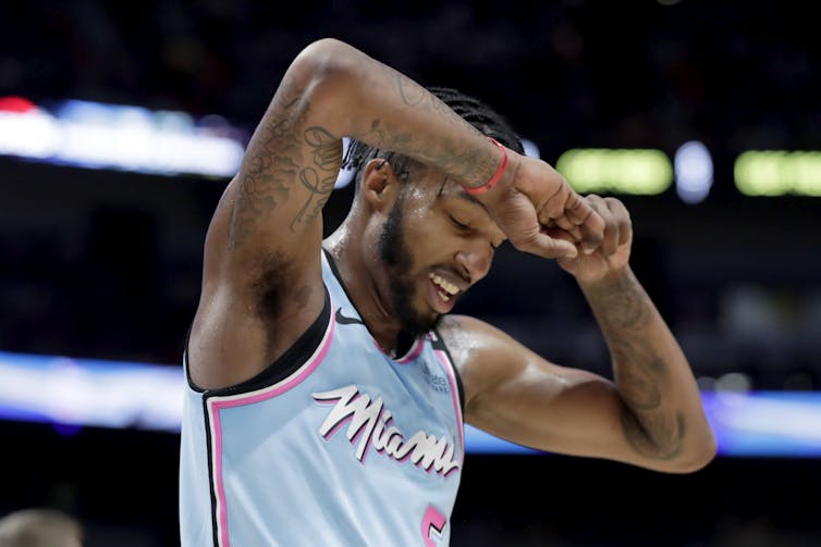 A Miami Heat player in a pale blue Miami Heat jersey looks down at the floor, his hands clasped against his forehead.
