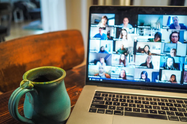 Several faces are shown on a laptop screen with a blue mug of coffee in the foreground.