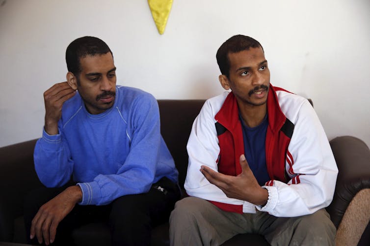 Two men, one gesturing with his hands, speak while sitting side by side on a dark-coloured couch.