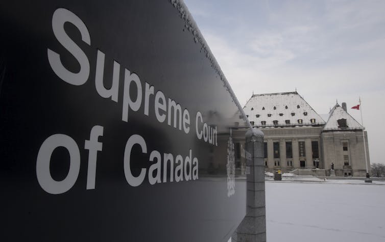 A sign reads Supreme Court of Canada with the Supreme Court buildings in the background with snow on the ground.