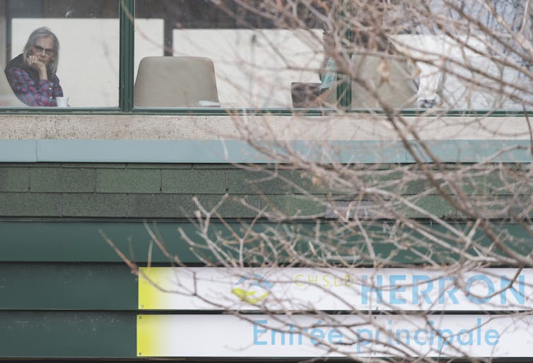 An elderly woman sits alone with her chin resting on her hand, looking out a window.