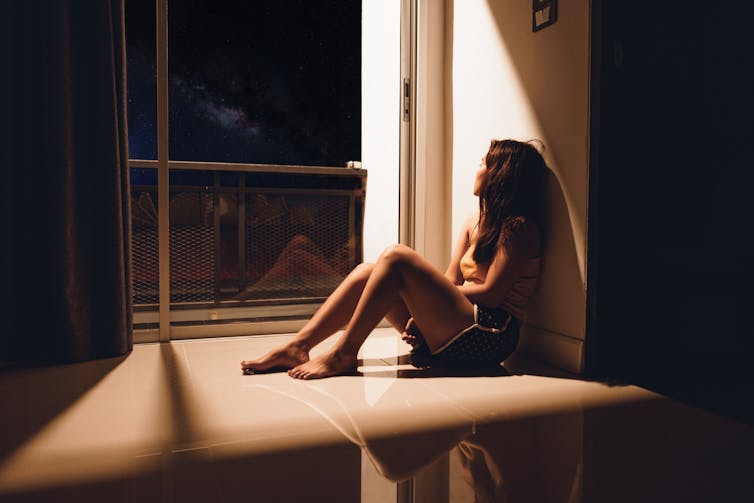 A young woman sits against the wall at home looking outside into the night.