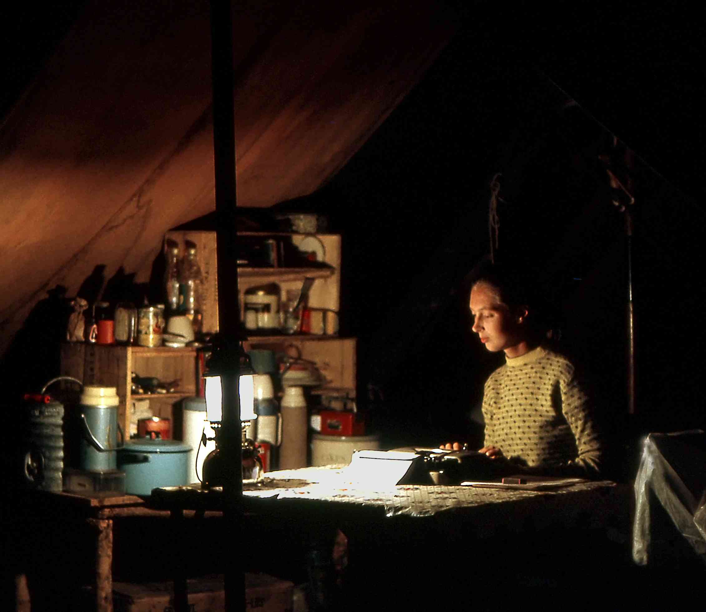 A young Jane Goodall writes notes on her desk in a tent by the dim light of a small lamp.