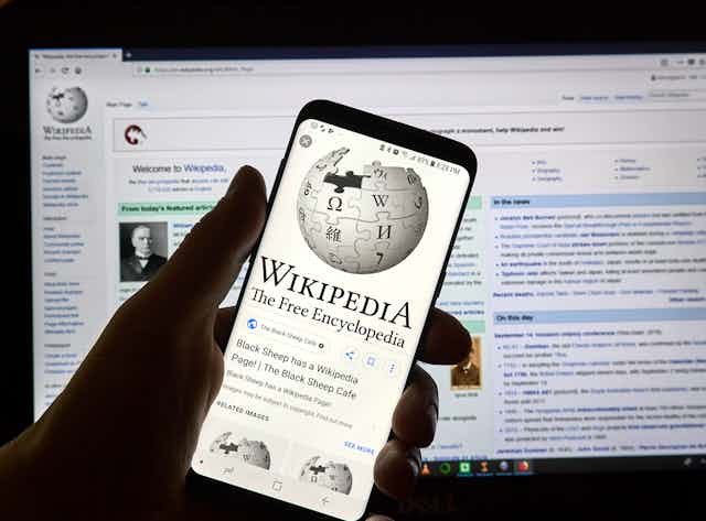 Una mano sujeta un móvil con la página de la Wikipedia. De fondo, página Wikipedia en pantalla de ordenador