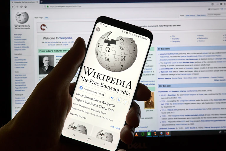 Una mano sujeta un móvil con la página de la Wikipedia. De fondo, página Wikipedia en pantalla de ordenador