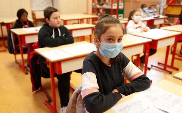 A school girl, her hair pulled back, wears a mask in a classroom. Classmates are seated behind her, some wearing masks and some bare-faced.