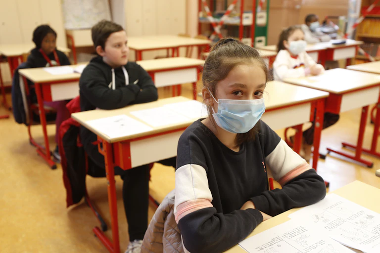 A school girl, her hair pulled back, wears a mask in a classroom. Classmates are seated behind her, some wearing masks and some bare-faced.