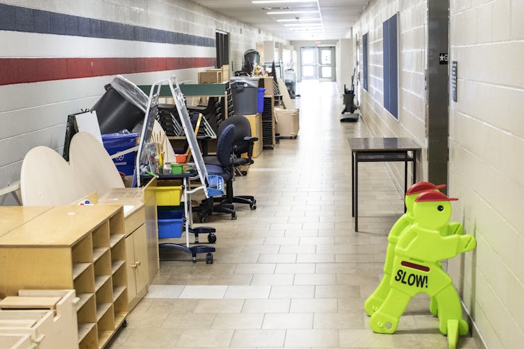 Desks, chairs and shelving units are pushed against the wall of an empty corridor in a school.