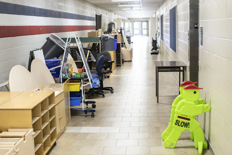 Desks, chairs and shelving units are pushed against the wall of an empty corridor in a school.