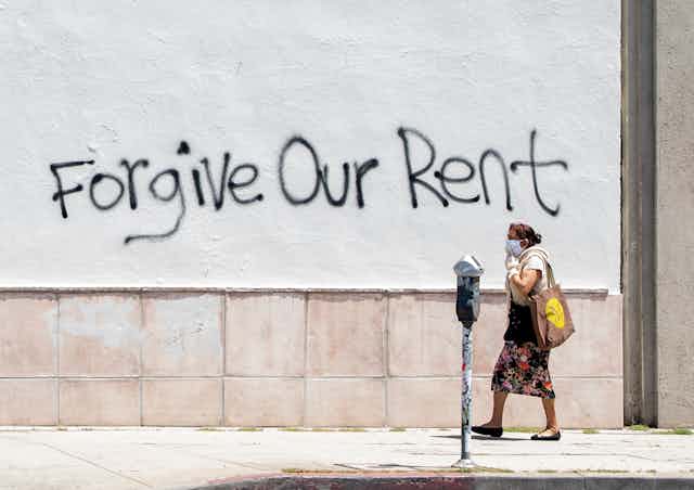 A woman wearing a mask walks past a wall bearing graffiti that reads "forgive our rent" on La Brea Ave in Los Angeles, California. 