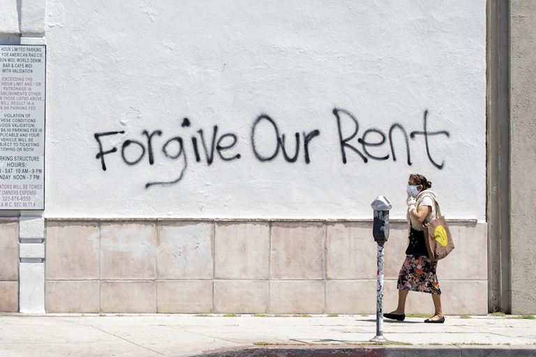 A woman wearing a mask walks past a wall bearing graffiti that reads "forgive our rent" on La Brea Ave in Los Angeles, California.