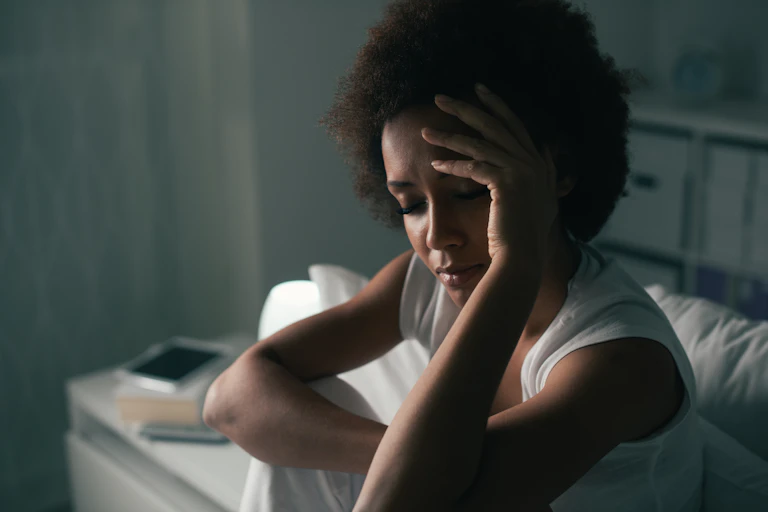 A Black woman sitting down with her eyes closed and her left hand on her forehead.