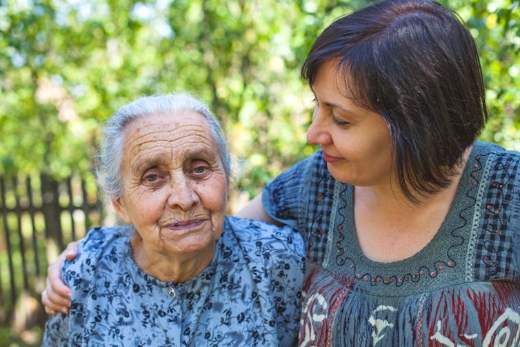 A middle-aged woman in a blue dress has her arm around and is looking at her older mother, also wearing a patterned blue dress.