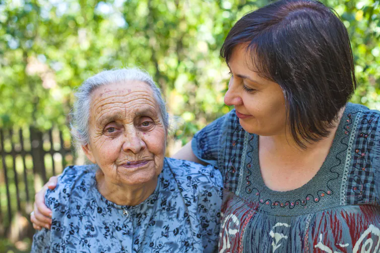 A middle-aged woman in a blue dress has her arm around and is looking at her older mother, also wearing a patterned blue dress.