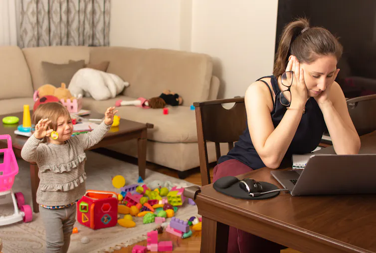 A white woman sits at a table in front of a laptop with her head in her hands as a toddler tries to get her attention.
