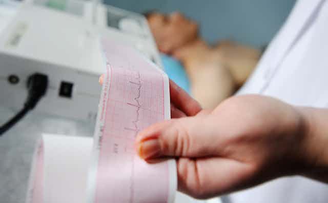 A doctor checks an ECG readout while a patient lies in the background