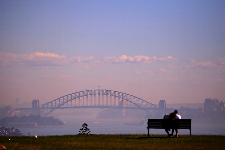 A couple on a bench look at the Sydney Harbour Bridge, shrouded in smoke.