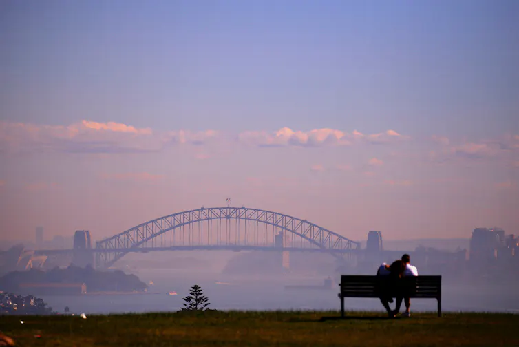 A couple on a bench look at the Sydney Harbour Bridge, shrouded in smoke.
