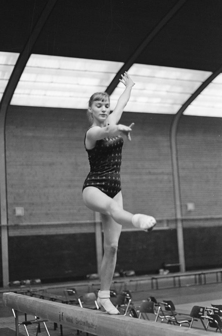 A teenage girl wears a leotard and stands on a beam.