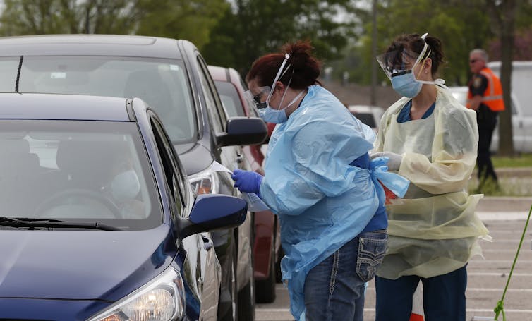 Two health care works standing outside preparing to perform a swab test at a drive-through testing station.
