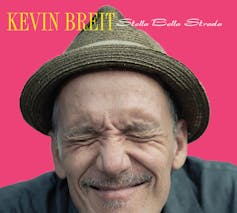 Kevin Breit wears a fedora and squints his eyes closed, while smiling against a fuschia background.