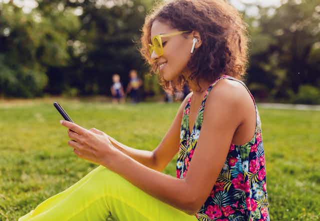 A woman listens and looks at a smartphone while wearing earbuds and sitting in the grass in summer.