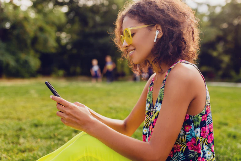 A woman listens and looks at a smartphone while wearing earbuds and sitting in the grass in summer.