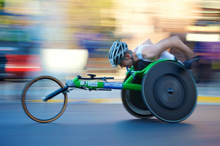 A disabled athlete wearing a helmet is seen, his head bowed, racing in his bright green racing wheelchair.