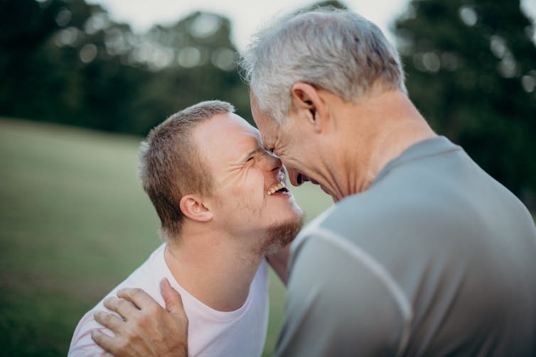 A disabled man smiles as he rubs noses with his father in a park.