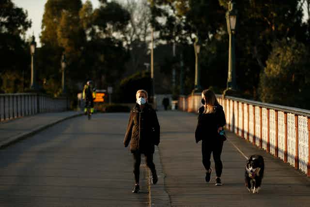 Two women wearing masks walk along a bridge. One has a dog.