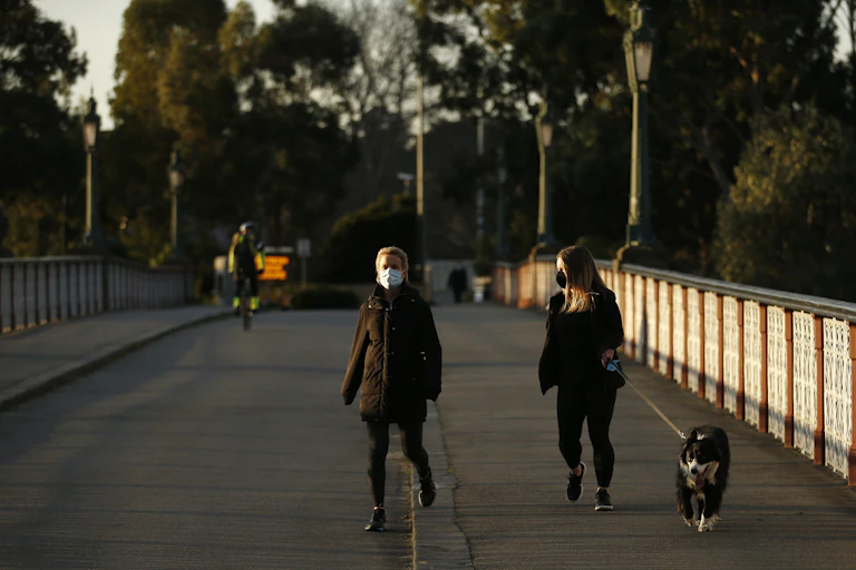 Two women wearing masks walk along a bridge. One has a dog.